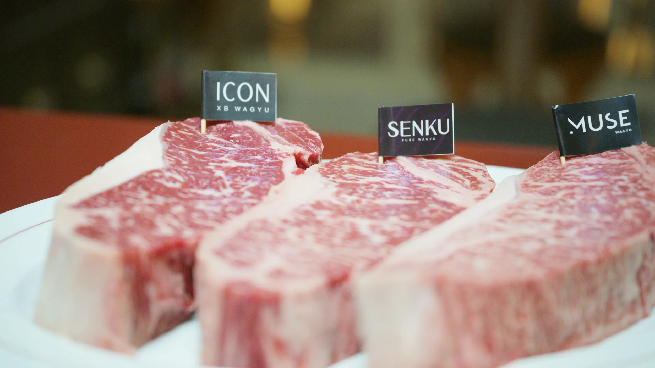 Three raw marbled striploins with ICON, SENKU, and MUSE flags on a red table