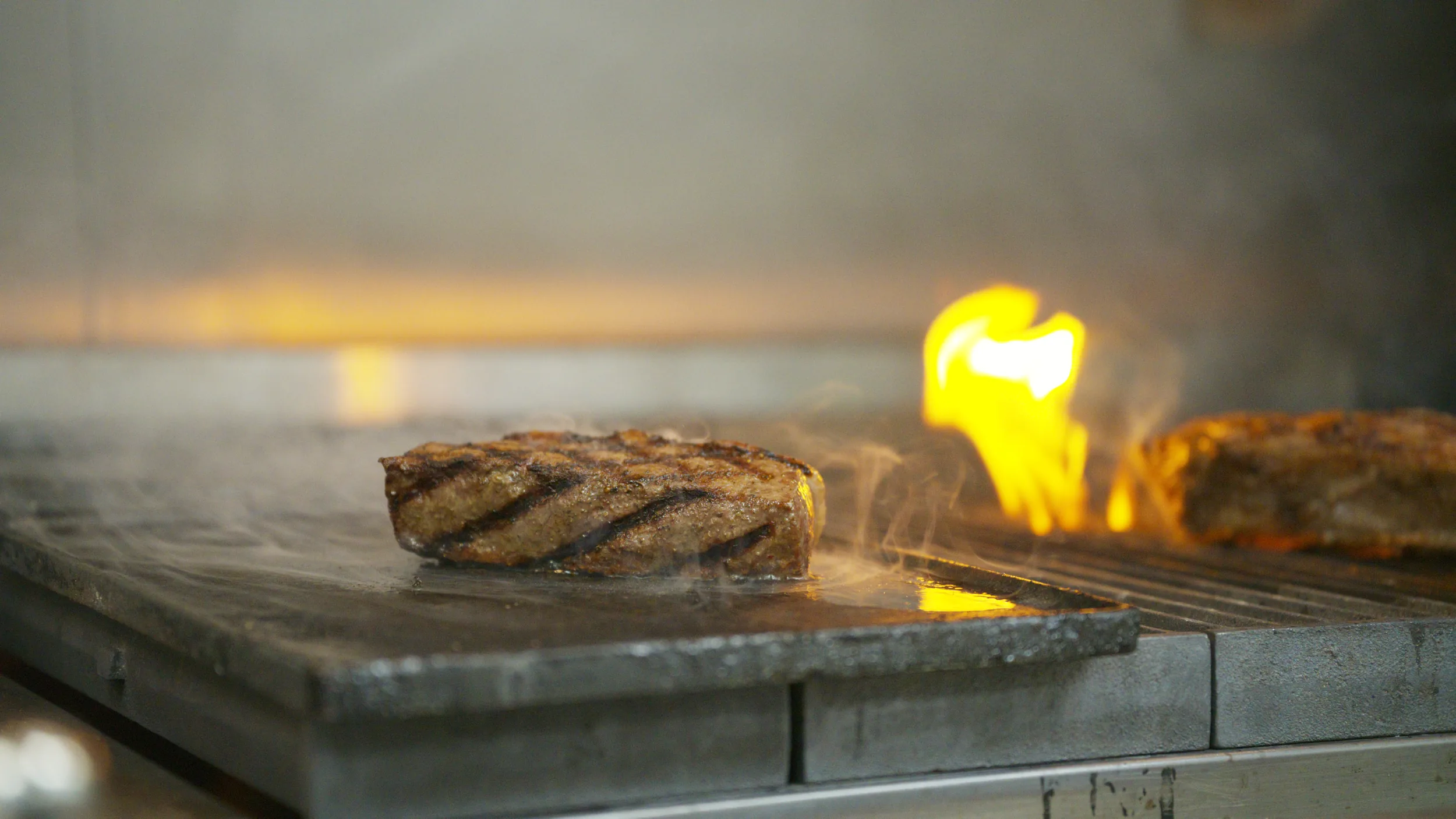 Grill-marked steak on a flat-top with a flame flaring behind it