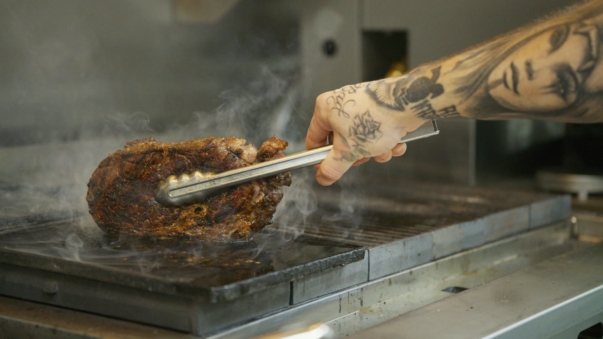 Tattooed forearm searing a steak's fat cap with tongs in smoke