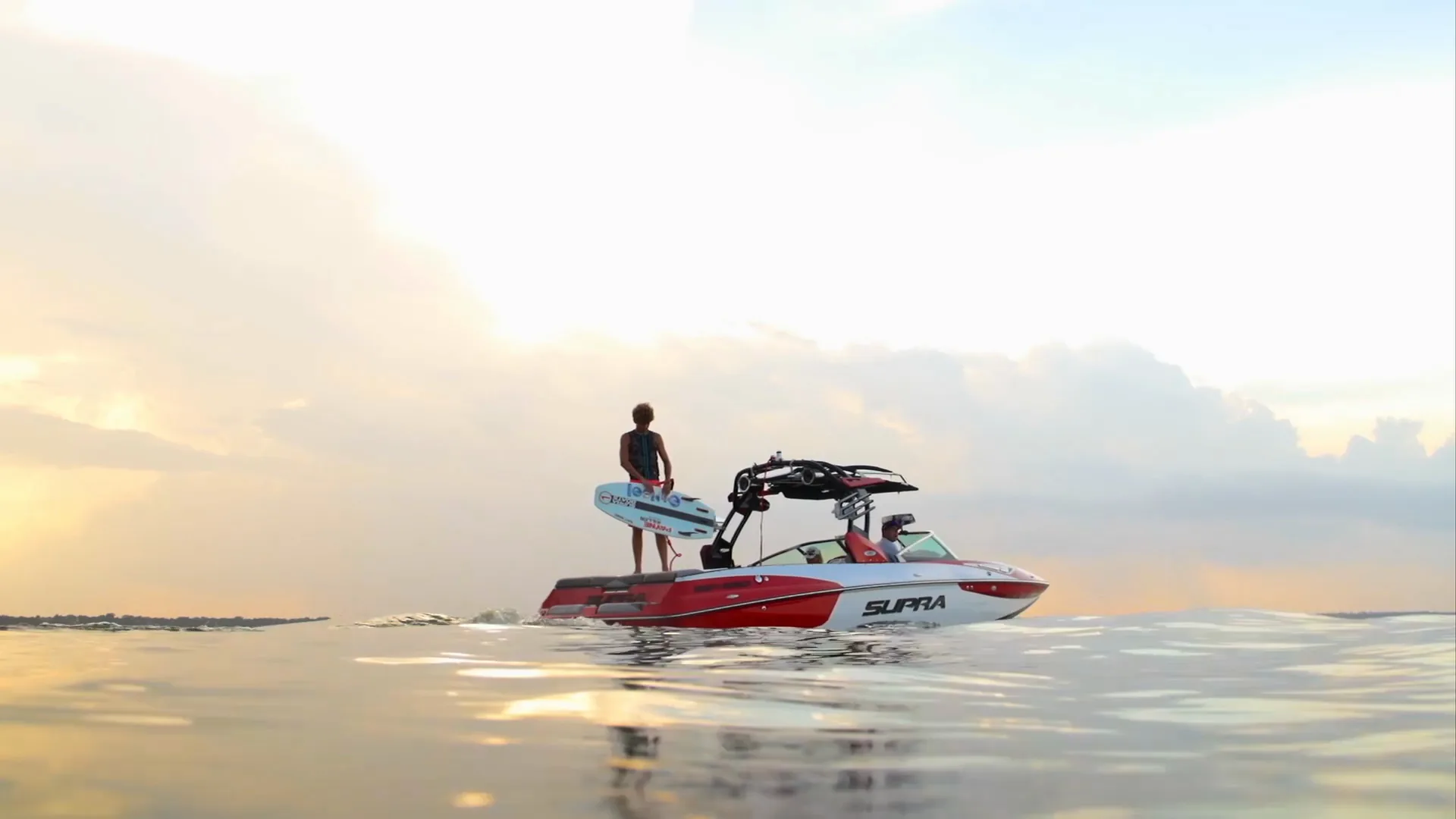Parker Payne standing on the Supra's deck with his board at sunset, shot from the water
