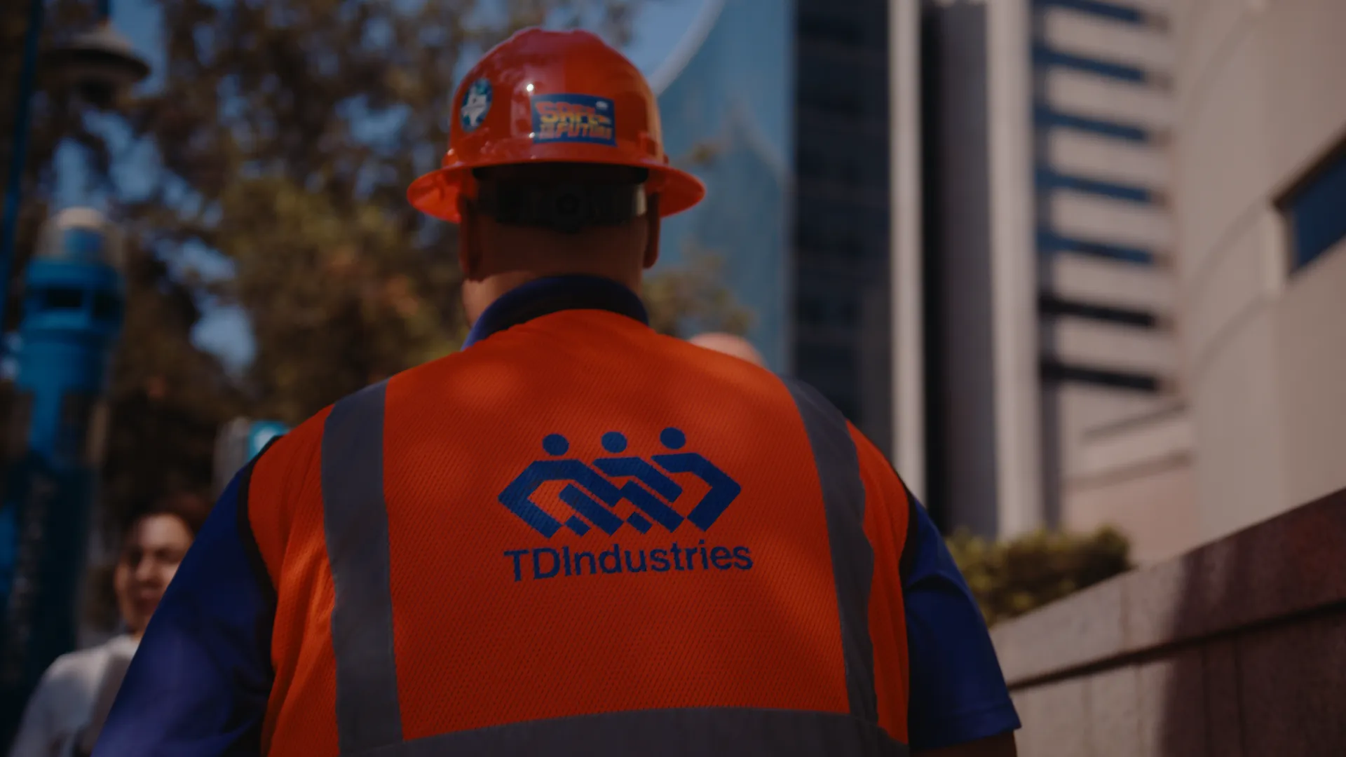 A worker in a TDIndustries safety vest and hard hat looking up at a downtown tower