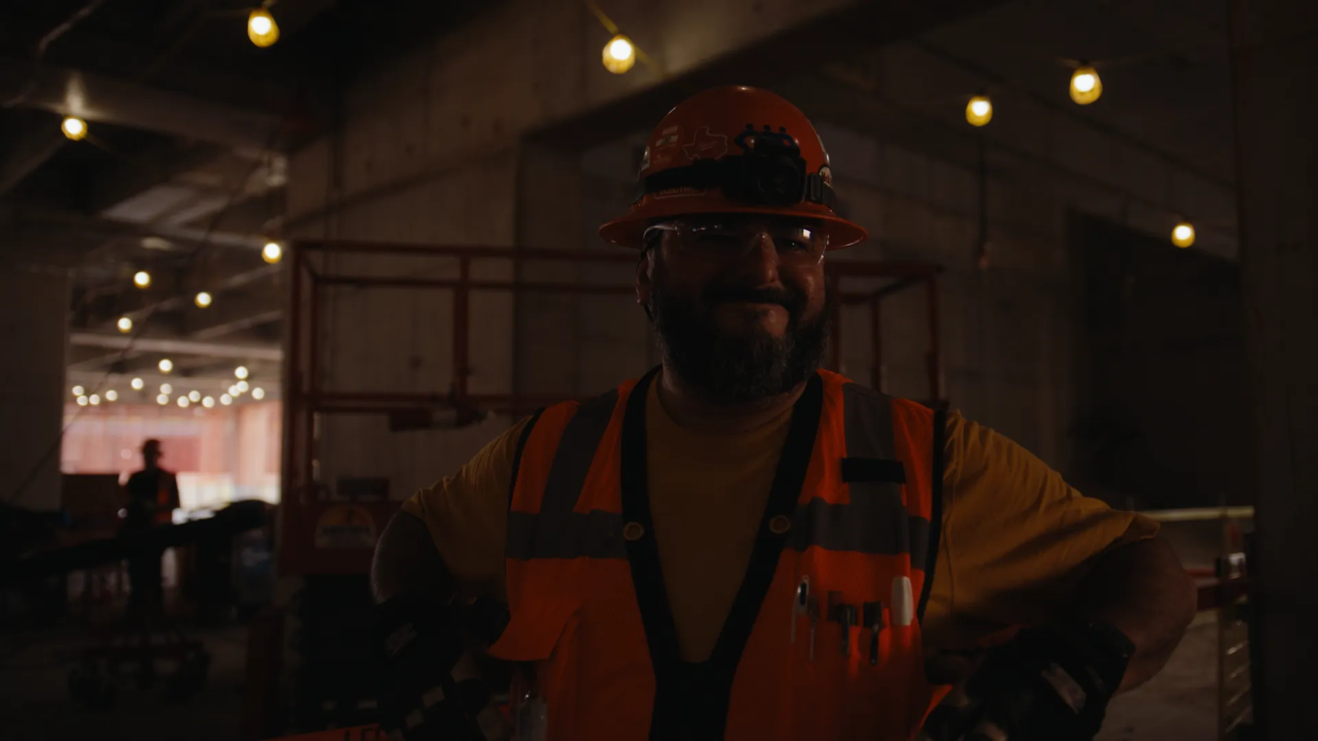 A smiling worker in a hard hat and vest beside a lift