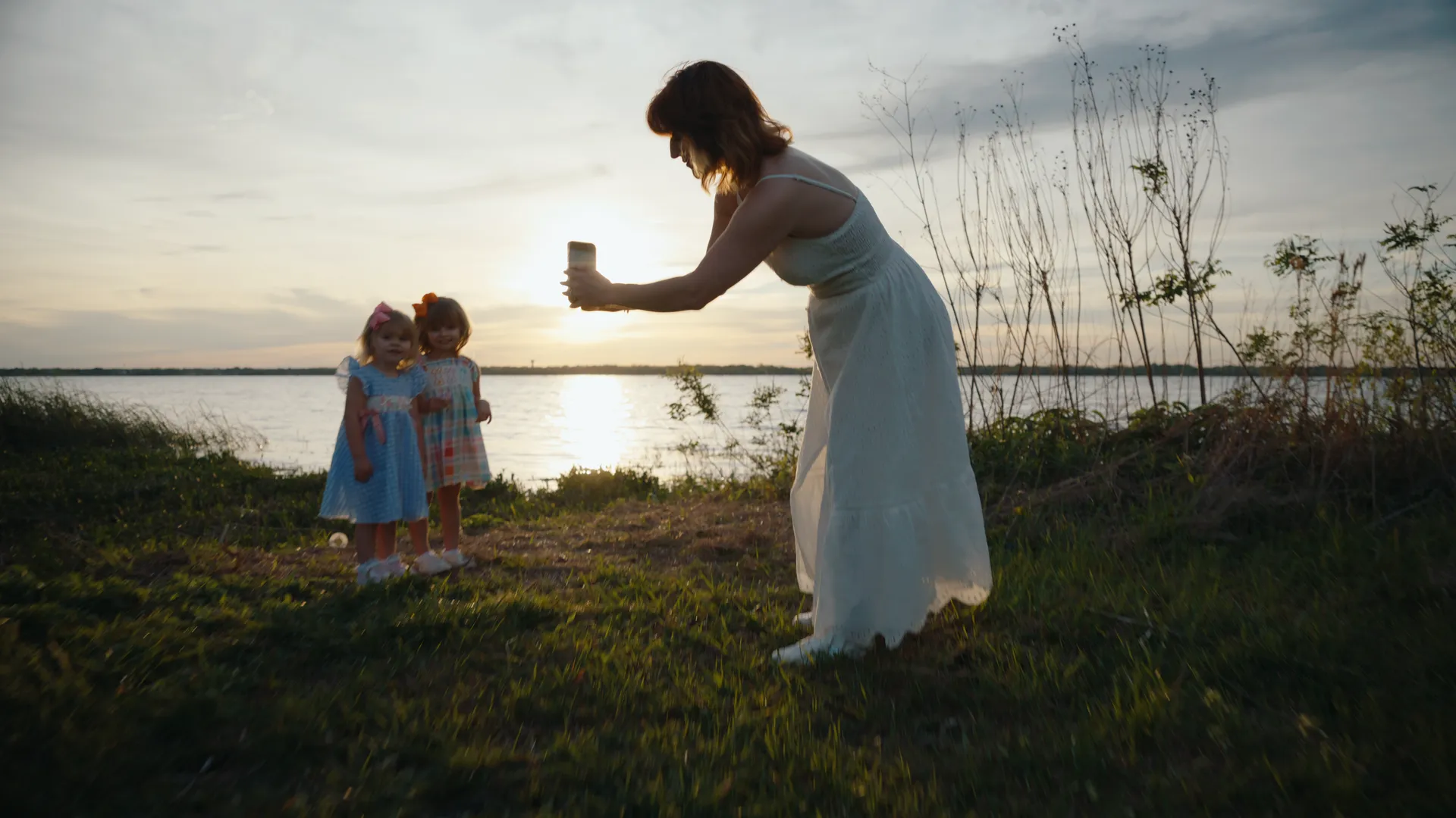 A woman in a white dress photographing two little girls on a lakeshore at sunset