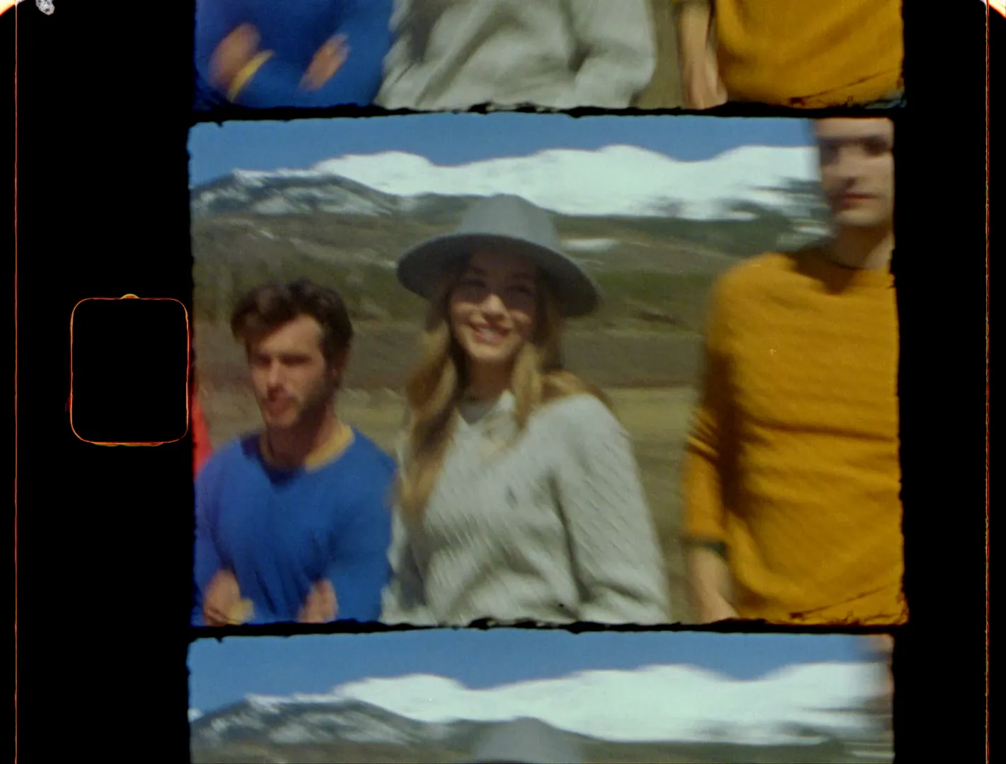Friends in blue, gray, and mustard sweaters walking under snowy peaks, one in a wide-brim hat
