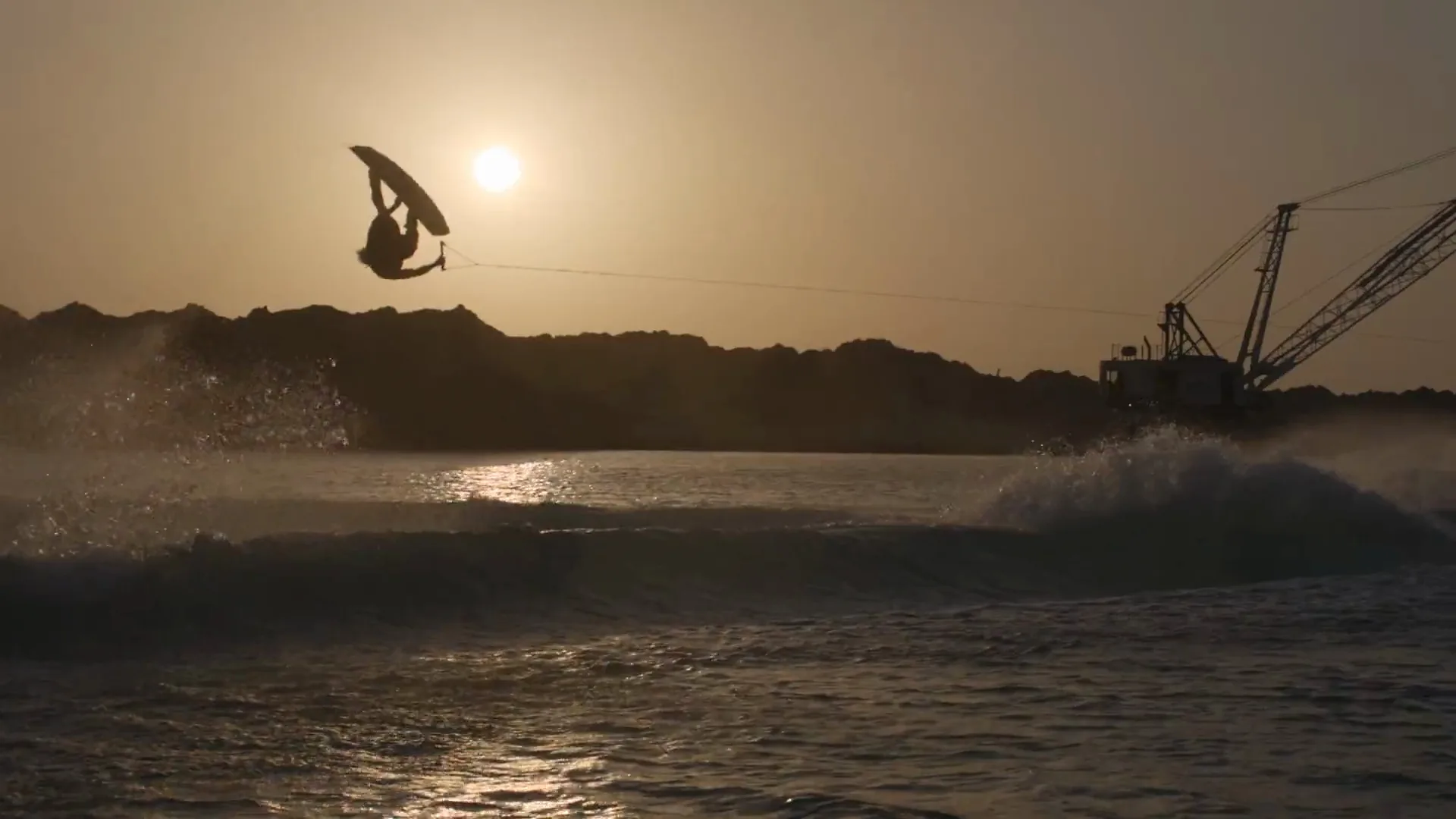 Mike Dowdy upside down grabbing his board at dusk, silhouetted against the low sun with a dragline crane on the shore